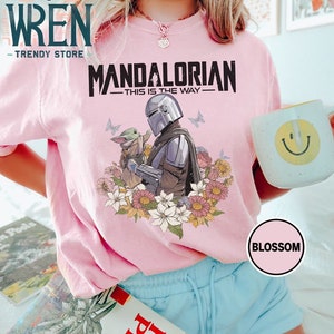 May include: Pink t-shirt featuring a graphic of the Mandalorian and Grogu from the Star Wars series. The graphic is surrounded by flowers. The text "Mandalorian - This is the Way -" is printed on the shirt.