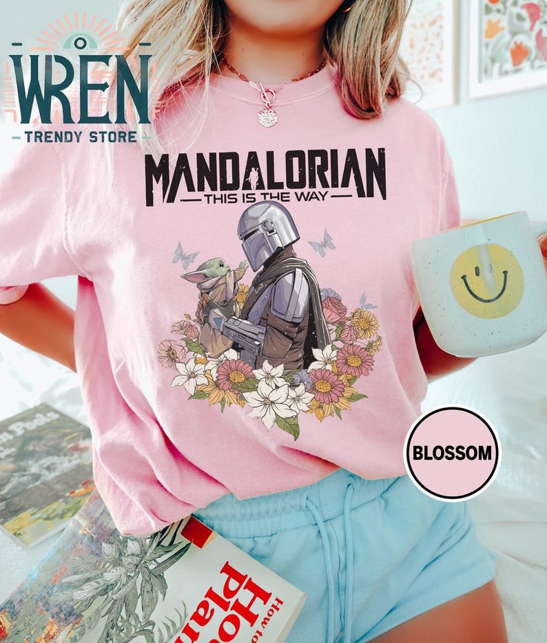 May include: Pink t-shirt featuring a graphic of the Mandalorian and Grogu from the Star Wars series. The graphic is surrounded by flowers. The text "Mandalorian - This is the Way -" is printed on the shirt.