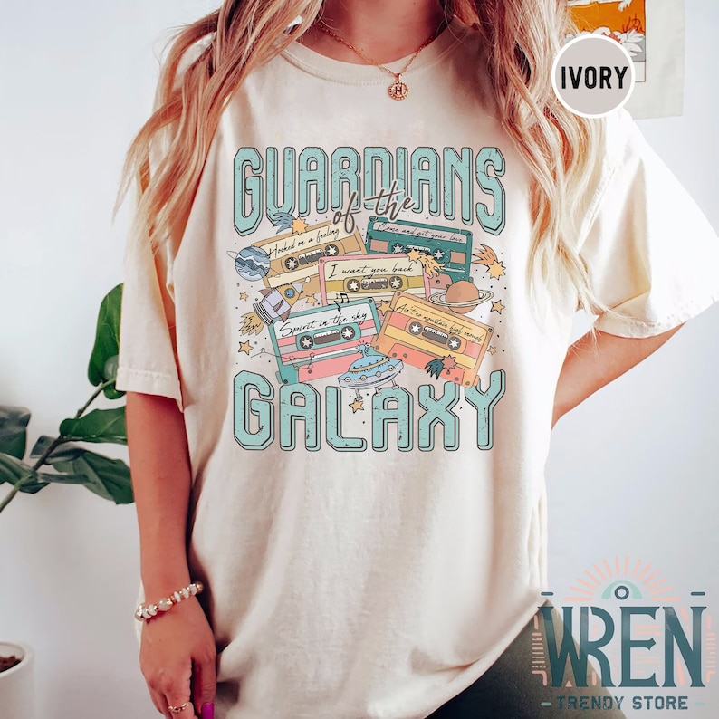 K&ouml;nnte beinhalten: Ein hellbeiges T-Shirt mit einem grafischen Design aus Kassetten und Planeten. Der Text "Guardians of the Galaxy" ist in blauen und gr&uuml;nen Buchstaben gedruckt.
