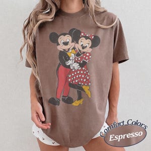 May include: A brown short-sleeved t-shirt featuring a graphic print of Mickey and Minnie Mouse embracing. Mickey wears a black jacket with red pants and a yellow bow tie, while Minnie wears a red dress with white polka dots and yellow shoes.