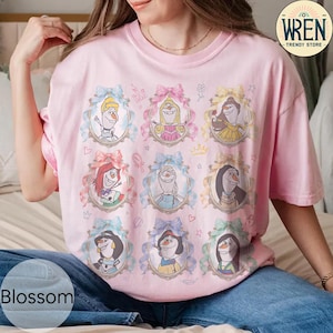 Puede incluir: Una camiseta rosa claro con nueve retratos enmarcados de personajes de dibujos animados, cada uno adornado con un lazo. Los retratos est&aacute;n dispuestos en tres filas de tres. La camiseta tiene mangas cortas y cuello redondo. La palabra "Blossom" es visible en la parte inferior izquierda.