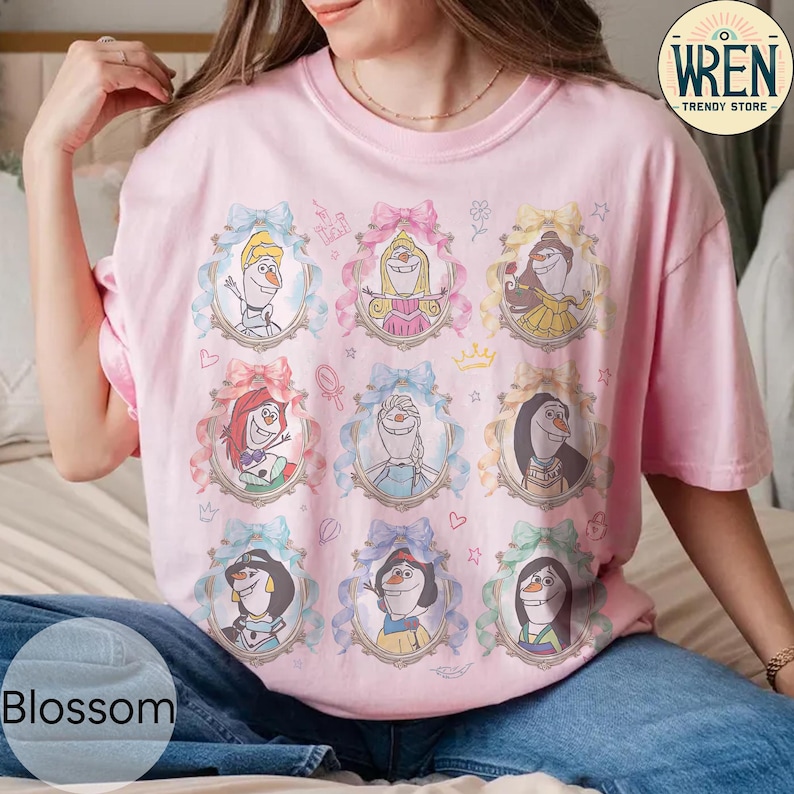 Puede incluir: Una camiseta rosa claro con nueve retratos enmarcados de personajes de dibujos animados, cada uno adornado con un lazo. Los retratos est&aacute;n dispuestos en tres filas de tres. La camiseta tiene mangas cortas y cuello redondo. La palabra "Blossom" es visible en la parte inferior izquierda.