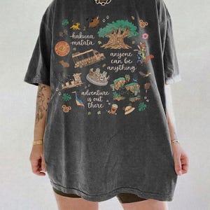 May include: Dark gray t-shirt with a colorful graphic featuring the text "hakuna matata", "anyone can be anything", and "adventure is out there". The graphic includes illustrations of a tree, animals, and a safari vehicle.