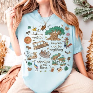 May include: Light blue t-shirt with a colorful graphic featuring the words "hakuna matata" and "adventure is out there." The design includes cartoon characters, a tree, and other themed illustrations.