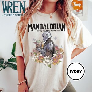 May include: A white t-shirt featuring a graphic print of the Mandalorian and Grogu, with the text "Mandalorian - This is the Way -" above them. The graphic is surrounded by flowers. The shirt is labelled "Ivory".