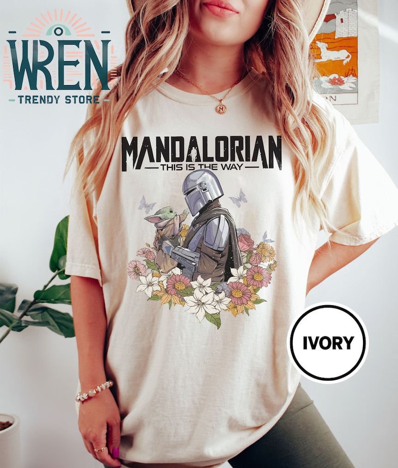 May include: A white t-shirt featuring a graphic print of the Mandalorian and Grogu, with the text "Mandalorian - This is the Way -" above them. The graphic is surrounded by flowers. The shirt is labelled "Ivory".