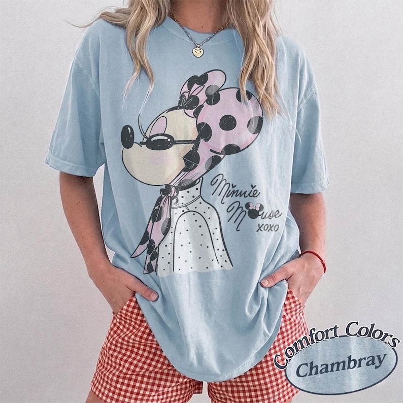 May include: Light blue t-shirt featuring a Minnie Mouse graphic. The graphic shows Minnie wearing sunglasses and a polka dot scarf. The text "Minnie Mouse xoxo" is below the graphic. The shirt is a Comfort Colors Chambray.