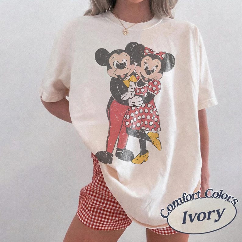May include: An ivory-colored short-sleeve t-shirt featuring a distressed graphic print of Mickey and Minnie Mouse embracing. The design shows Mickey in red pants and a yellow bowtie, while Minnie wears a red and white polka dot dress and yellow shoes.