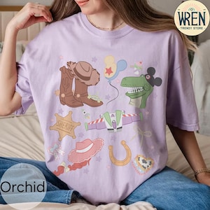 May include: A light purple t-shirt featuring cartoon illustrations of a cowboy hat, boots, a sheriff badge, a dinosaur, a space ship, a horseshoe, and other elements. The shirt has the text "WREN TRENDY STORE" on the upper right corner and "Orchid" on the bottom left.