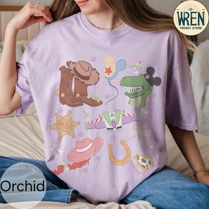 May include: A light purple t-shirt featuring cartoon illustrations of a cowboy hat, boots, a sheriff badge, a dinosaur, a space ship, a horseshoe, and other elements. The shirt has the text "WREN TRENDY STORE" on the upper right corner and "Orchid" on the bottom left.