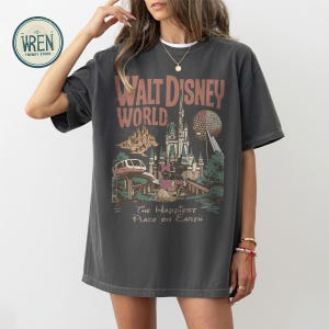Może przedstawiać: Ciemnoszara koszulka z grafiką w stylu vintage z Walt Disney World. Projekt zawiera nazwę parku, zamek, Spaceship Earth i frazę "The Happiest Place on Earth".