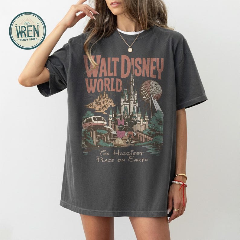 Może przedstawiać: Ciemnoszara koszulka z grafiką w stylu vintage z Walt Disney World. Projekt zawiera nazwę parku, zamek, Spaceship Earth i frazę "The Happiest Place on Earth".