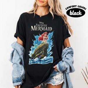 May include: A black t-shirt featuring a graphic of Ariel from The Little Mermaid sitting on a rock amidst blue ocean waves. The text Disney The Little Mermaid is printed above the character. A Comfort Colors black label is visible in the corner.