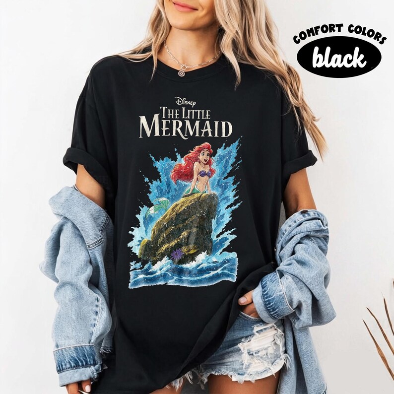May include: A black t-shirt featuring a graphic of Ariel from The Little Mermaid sitting on a rock amidst blue ocean waves. The text Disney The Little Mermaid is printed above the character. A Comfort Colors black label is visible in the corner.