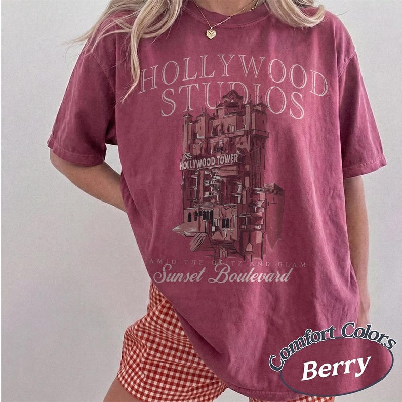 May include: A berry-colored short-sleeve t-shirt featuring a graphic of the Hollywood Tower Hotel. The text reads Hollywood Studios, The Hollywood Tower, and Sunset Boulevard. The shirt has a crew neckline and a relaxed, loose-fitting silhouette.