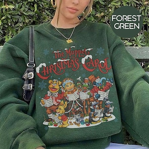 May include: Forest green sweatshirt featuring a graphic from "The Muppet Christmas Carol." The design includes the title in red and white, with illustrations of Muppet characters. The sweatshirt is a solid forest green color.