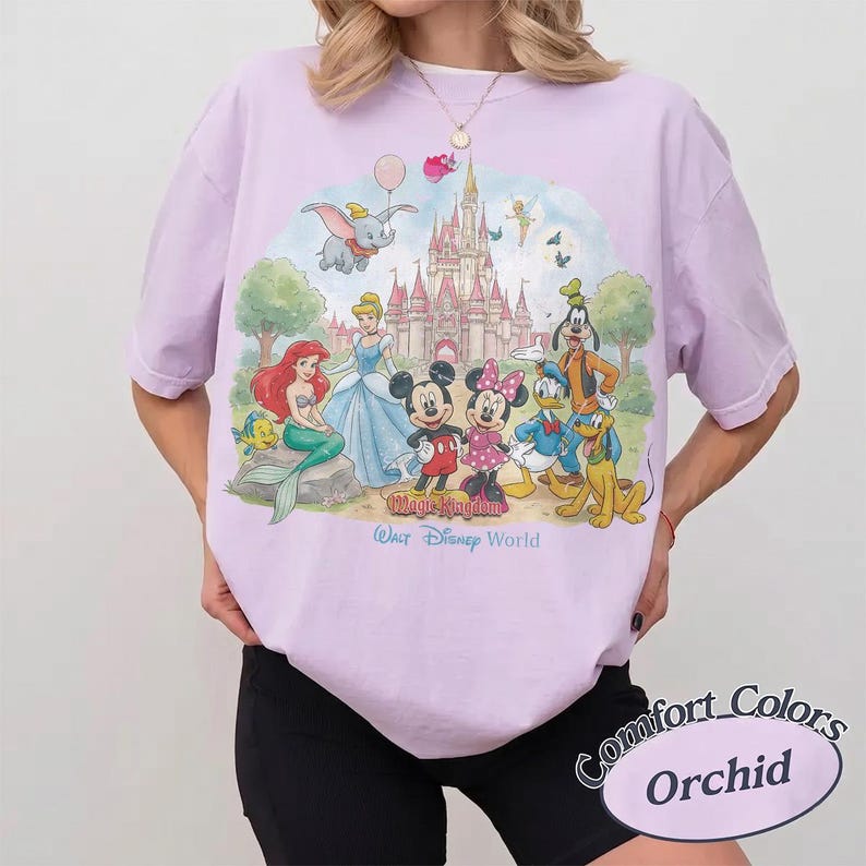 May include: A light purple t-shirt featuring a colorful graphic of Disney characters including Mickey Mouse, Minnie Mouse, Ariel, Cinderella, Dumbo, and Goofy in front of a castle. The text reads Magic Kingdom and Walt Disney World.