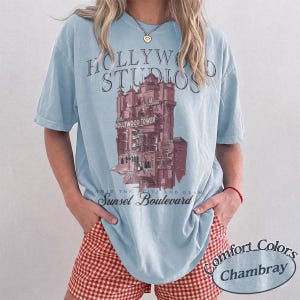 May include: A light blue short-sleeve t-shirt featuring a graphic of the Hollywood Tower Hotel. The text reads Hollywood Studios, The Hollywood Tower, and Sunset Boulevard. The shirt is made by Comfort Colors in the color Chambray.