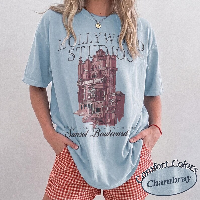 May include: A light blue short-sleeve t-shirt featuring a graphic of the Hollywood Tower Hotel. The text reads Hollywood Studios, The Hollywood Tower, and Sunset Boulevard. The shirt is made by Comfort Colors in the color Chambray.