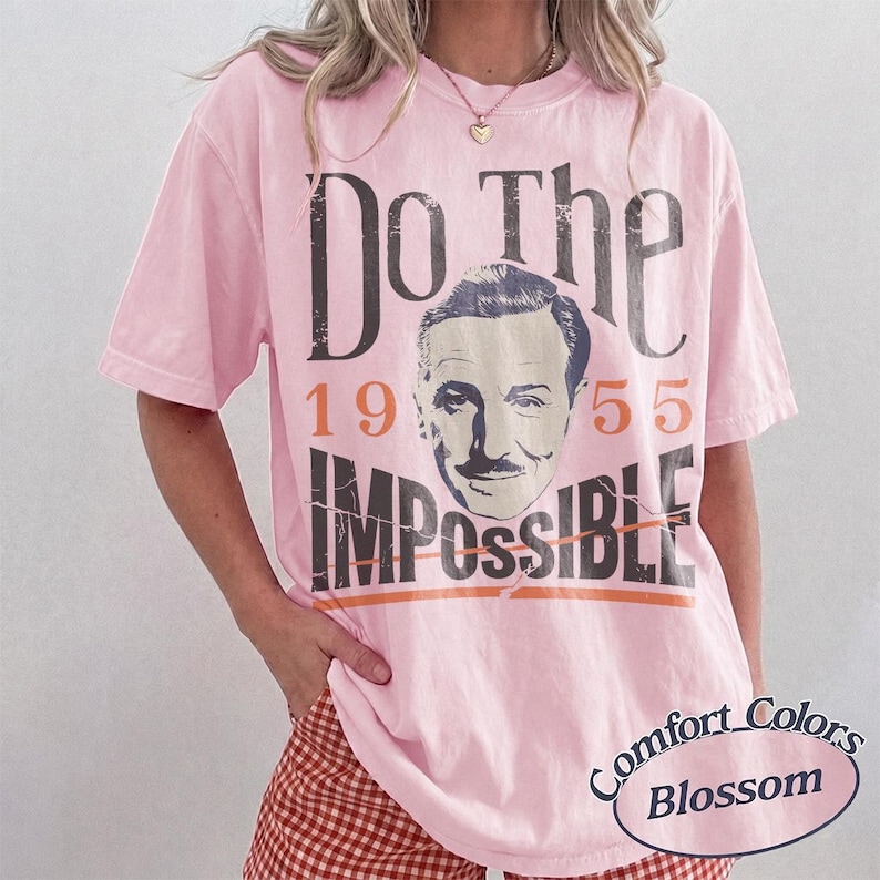 May include: Pink short-sleeved t-shirt with the text "Do The Impossible" and a vintage portrait. The year "1955" is printed above the word "Impossible". The shirt is labeled "Comfort Colors Blossom".