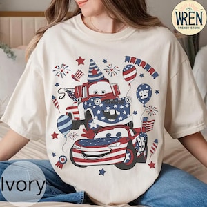 Puede incluir: Camiseta de color marfil con un dise&ntilde;o patri&oacute;tico con coches de dibujos animados, fuegos artificiales y globos en rojo, blanco y azul. El dise&ntilde;o incluye el n&uacute;mero "95" y estrellas, con el texto "WREN TRENDY STORE" en la parte superior derecha.