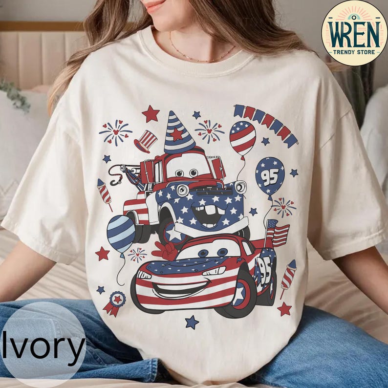 Puede incluir: Camiseta de color marfil con un dise&ntilde;o patri&oacute;tico con coches de dibujos animados, fuegos artificiales y globos en rojo, blanco y azul. El dise&ntilde;o incluye el n&uacute;mero "95" y estrellas, con el texto "WREN TRENDY STORE" en la parte superior derecha.