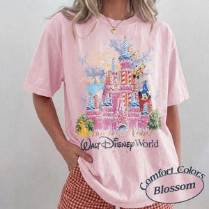 Może przedstawiać: Jasnor&oacute;żowa koszulka z grafiką zamku Kopciuszka, Myszki Miki i innych postaci. Pod spodem nadrukowano napis "This is Now! Walt Disney World 1997". Koszulka oznaczona jest jako "Comfort Colors Blossom".