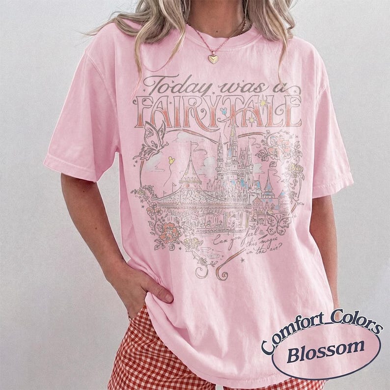 Puede incluir: Una camiseta de manga corta de color rosa claro con un estampado gr&aacute;fico de un castillo, flores y una mariposa. El texto dice Today was a FAIRY TALE y Can you feel this magic in the air? La prenda tiene un cuello redondo.