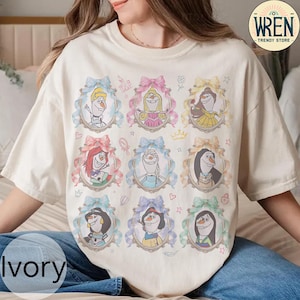 Puede incluir: Camiseta de color marfil con nueve retratos enmarcados de personajes animados, cada uno rodeado por un lazo de color pastel. Los retratos incluyen personajes con varios peinados y atuendos. La camiseta tiene cuello redondo y mangas cortas. El texto "Ivory" est&aacute; en la parte inferior.