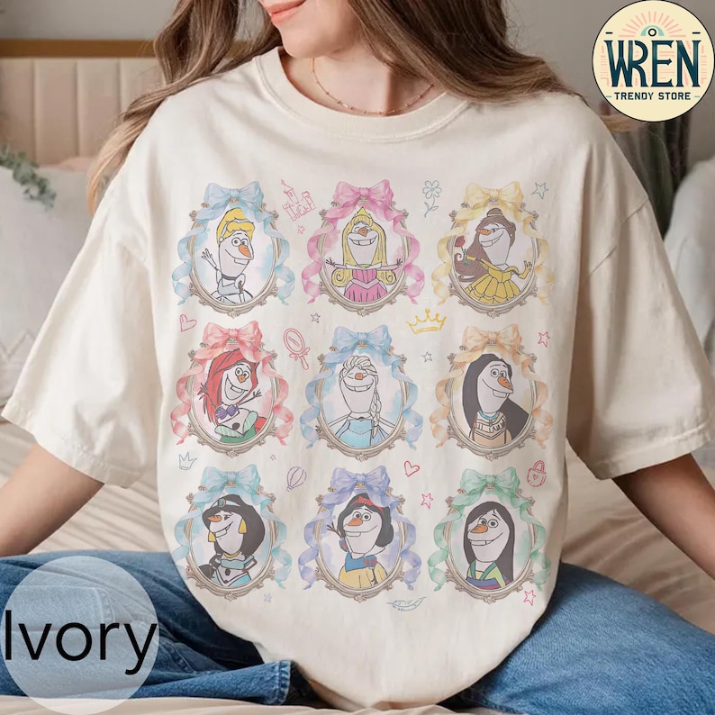 Puede incluir: Camiseta de color marfil con nueve retratos enmarcados de personajes animados, cada uno rodeado por un lazo de color pastel. Los retratos incluyen personajes con varios peinados y atuendos. La camiseta tiene cuello redondo y mangas cortas. El texto "Ivory" est&aacute; en la parte inferior.
