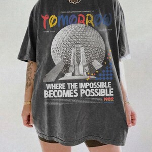 May include: A charcoal gray oversized t-shirt featuring a large graphic of a geodesic dome. The text reads "TOMORROW," "SPARK YOUR IMAGINATION," "WHERE THE IMPOSSIBLE BECOMES POSSIBLE," and "1982." The design includes small colorful geometric shapes.