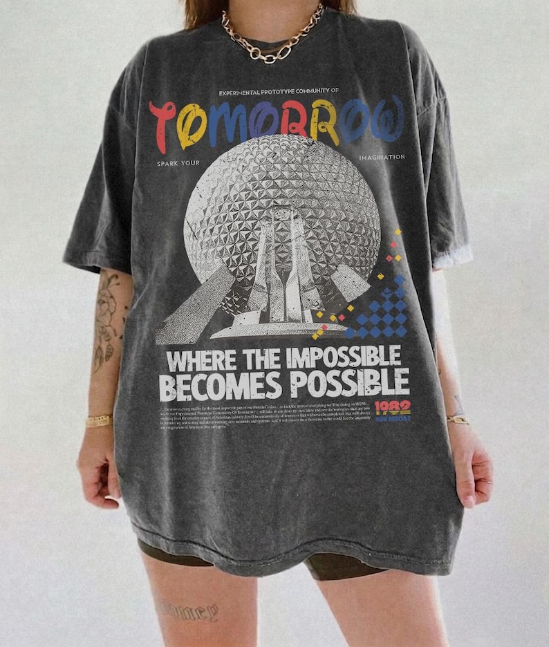 May include: A charcoal gray oversized t-shirt featuring a large graphic of a geodesic dome. The text reads "TOMORROW," "SPARK YOUR IMAGINATION," "WHERE THE IMPOSSIBLE BECOMES POSSIBLE," and "1982." The design includes small colorful geometric shapes.