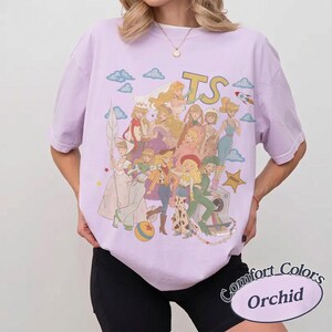 May include: A light purple short-sleeve t-shirt featuring a graphic print of various animated characters in a collage style. The design includes the letters TS, a small rocket, clouds, and a yellow star badge. The shirt has a crew neckline.