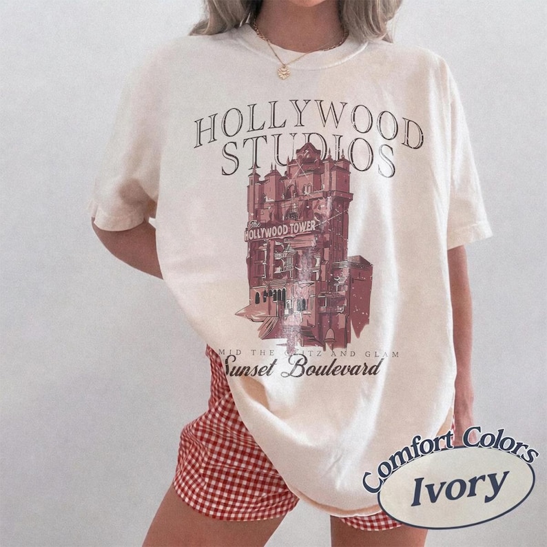 May include: An ivory short-sleeve t-shirt featuring a graphic of the Hollywood Tower Hotel. The text reads HOLLYWOOD STUDIOS at the top and Sunset Boulevard at the bottom. The design is printed in shades of maroon and dark brown on the front.