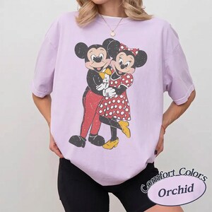 May include: A light purple short-sleeve t-shirt featuring a distressed graphic print of Mickey and Minnie Mouse embracing. Mickey wears red pants and a yellow bowtie, while Minnie wears a red and white polka dot dress and yellow shoes.