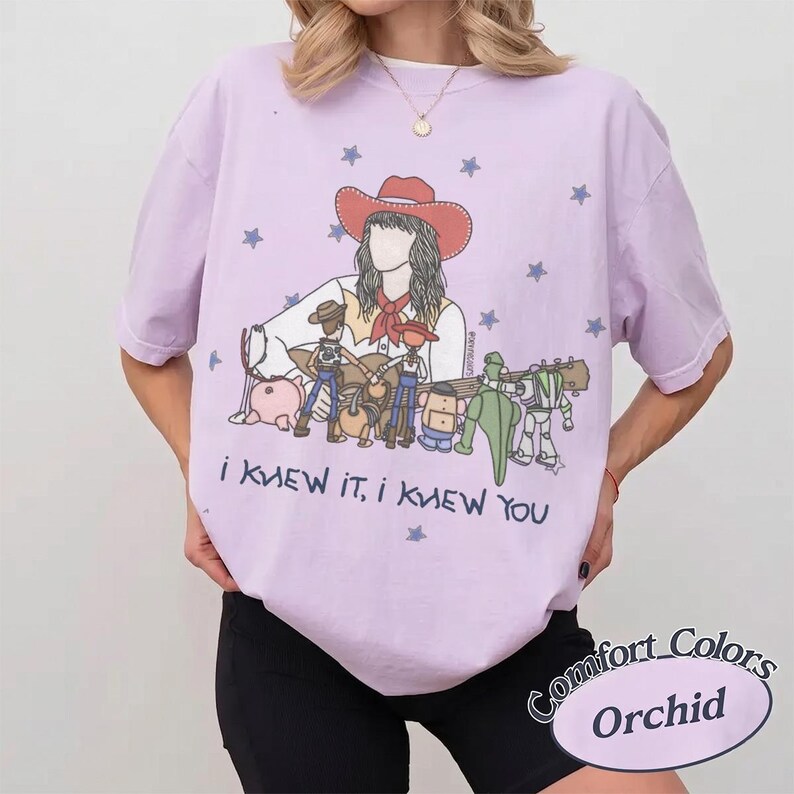 May include: A light purple t-shirt featuring a graphic of a person in a cowboy hat surrounded by various toy characters. The design includes blue stars and the text "I KNEW IT, I KNEW YOU" printed in dark blue lettering below the illustrated figures.