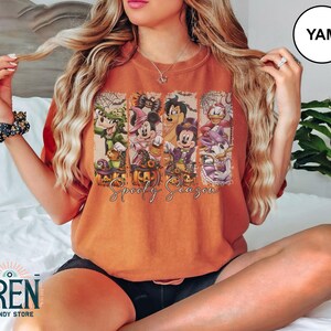 May include: Orange t-shirt featuring a Halloween-themed graphic with cartoon characters and pumpkins. The text "Spooky Season" is printed below the characters. The shirt is a comfortable fit.
