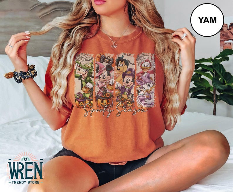 May include: Orange t-shirt featuring a Halloween-themed graphic with cartoon characters and pumpkins. The text "Spooky Season" is printed below the characters. The shirt is a comfortable fit.