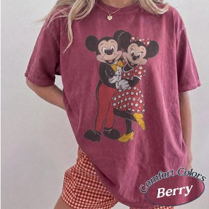 May include: A berry-colored short-sleeve t-shirt featuring a graphic print of Mickey and Minnie Mouse embracing. Mickey wears a black tuxedo with a yellow bowtie and red pants, while Minnie wears a red and white polka dot dress and yellow shoes.