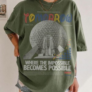 May include: An olive green t-shirt featuring a large graphic of a geodesic dome structure. The text reads "EXPERIMENTAL PROTOTYPE COMMUNITY OF TOMORROW", "SPARK YOUR IMAGINATION", and "WHERE THE IMPOSSIBLE BECOMES POSSIBLE" with small geometric shapes.