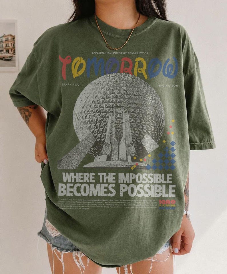 May include: An olive green t-shirt featuring a large graphic of a geodesic dome structure. The text reads "EXPERIMENTAL PROTOTYPE COMMUNITY OF TOMORROW", "SPARK YOUR IMAGINATION", and "WHERE THE IMPOSSIBLE BECOMES POSSIBLE" with small geometric shapes.