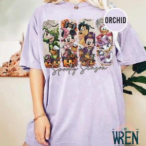 May include: A light purple t-shirt featuring a Halloween-themed graphic with cartoon characters and pumpkins. The graphic includes the text "Spooky Season" in a script font. The shirt has a relaxed fit and short sleeves.