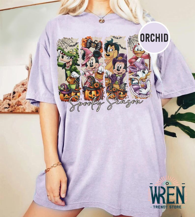May include: A light purple t-shirt featuring a Halloween-themed graphic with cartoon characters and pumpkins. The graphic includes the text "Spooky Season" in a script font. The shirt has a relaxed fit and short sleeves.