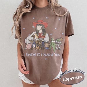 May include: A brown t-shirt featuring a graphic illustration of a person in a red cowboy hat playing a guitar, surrounded by various toy characters. The text "I KNEW IT, I KNEW YOU" is printed below the illustration, accented by small white stars.