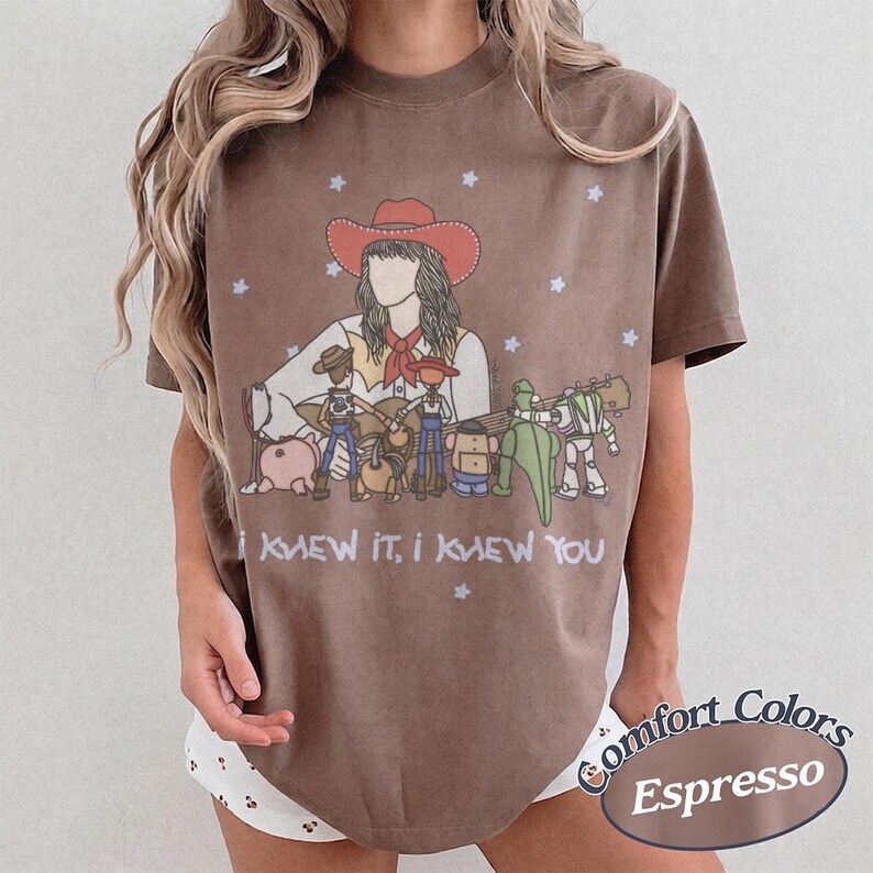 May include: A brown t-shirt featuring a graphic illustration of a person in a red cowboy hat playing a guitar, surrounded by various toy characters. The text "I KNEW IT, I KNEW YOU" is printed below the illustration, accented by small white stars.
