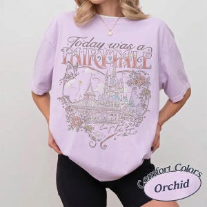 Puede incluir: Una camiseta de manga corta de color morado claro con un estampado gr&aacute;fico de un castillo, una mariposa y flores. El texto dice Today was a FAIRYTALE y Can you feel this magic in the air? La camiseta tiene un cuello redondo.