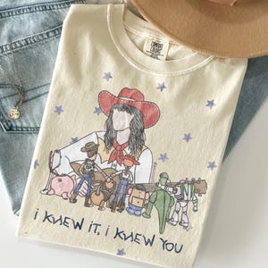 May include: A cream-coloured t-shirt featuring a graphic illustration of a person in a red cowboy hat surrounded by various toy characters and blue stars. The text "I KNEW IT, I KNEW YOU" is printed in blue at the bottom of the graphic design.