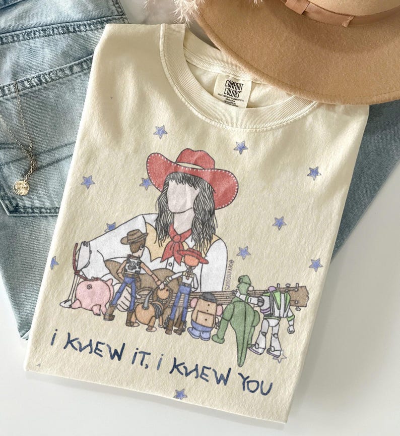 May include: A cream-coloured t-shirt featuring a graphic illustration of a person in a red cowboy hat surrounded by various toy characters and blue stars. The text "I KNEW IT, I KNEW YOU" is printed in blue at the bottom of the graphic design.