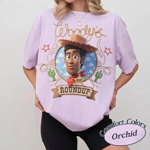 K&ouml;nnte beinhalten: Ein hellviolettes Kurzarm-T-Shirt mit einer Grafik der Figur Woody aus Toy Story in einem kreisf&ouml;rmigen Rahmen. Das Design enth&auml;lt den Text Woody's Roundup, einen Sheriff-Stern, Kakteen und Seilmotive in einem Vintage-inspirierten Stil.