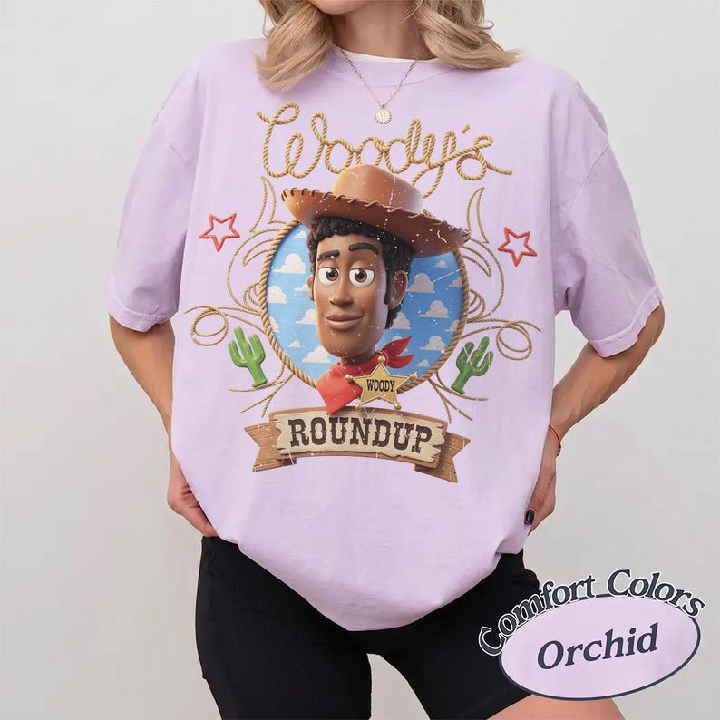 K&ouml;nnte beinhalten: Ein hellviolettes Kurzarm-T-Shirt mit einer Grafik der Figur Woody aus Toy Story in einem kreisf&ouml;rmigen Rahmen. Das Design enth&auml;lt den Text Woody's Roundup, einen Sheriff-Stern, Kakteen und Seilmotive in einem Vintage-inspirierten Stil.